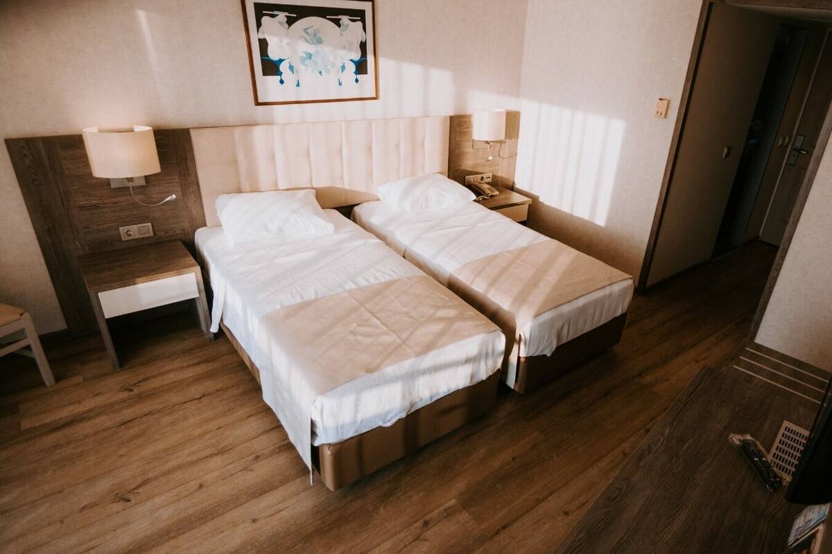 A hotel room with two beds, wooden flooring, bedside tables with lamps, a framed picture on the wall, and a door leading to another room.