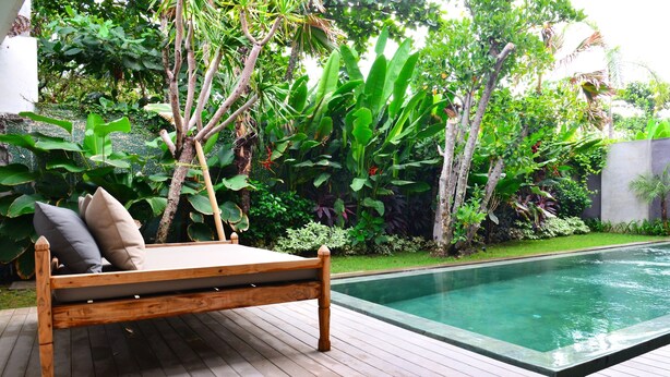 Une terrasse en bois avec un lit en bois et des coussins, entourée d’une végétation luxuriante et d’une piscine.
