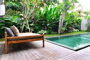 A wooden deck with a wooden bed and pillows, surrounded by lush greenery and a swimming pool. - Jabu Beach Villa (Canggu)