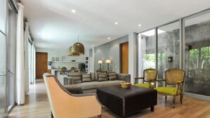 Un salon spacieux avec un canapé, des fauteuils et une table basse. Au fond de la pièce, on trouve un coin cuisine avec une table à manger et des chaises. La pièce est dotée de grandes fenêtres offrant une vue sur la verdure environnante.