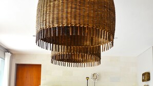 Un coin repas avec une grande table en bois, deux chaises en osier, un lustre suspendu en osier et, au fond, une cuisine.