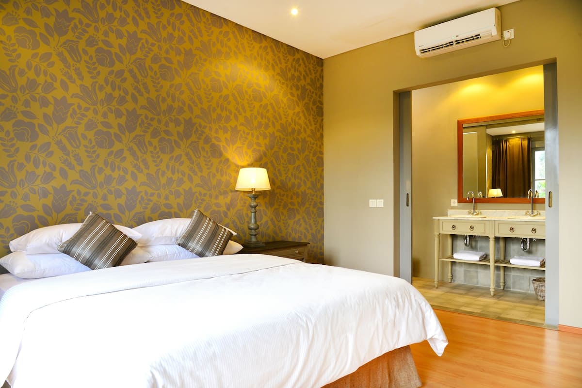 Habitación de hotel con cama, mesita de noche, lámpara, baño con espejo y aire acondicionado en la pared.