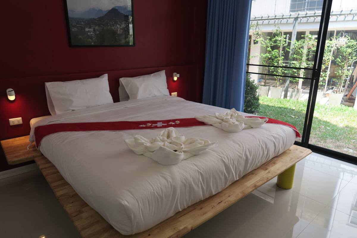 A modern hotel room with a large bed, wooden headboard, and a view of a green landscape through large windows.