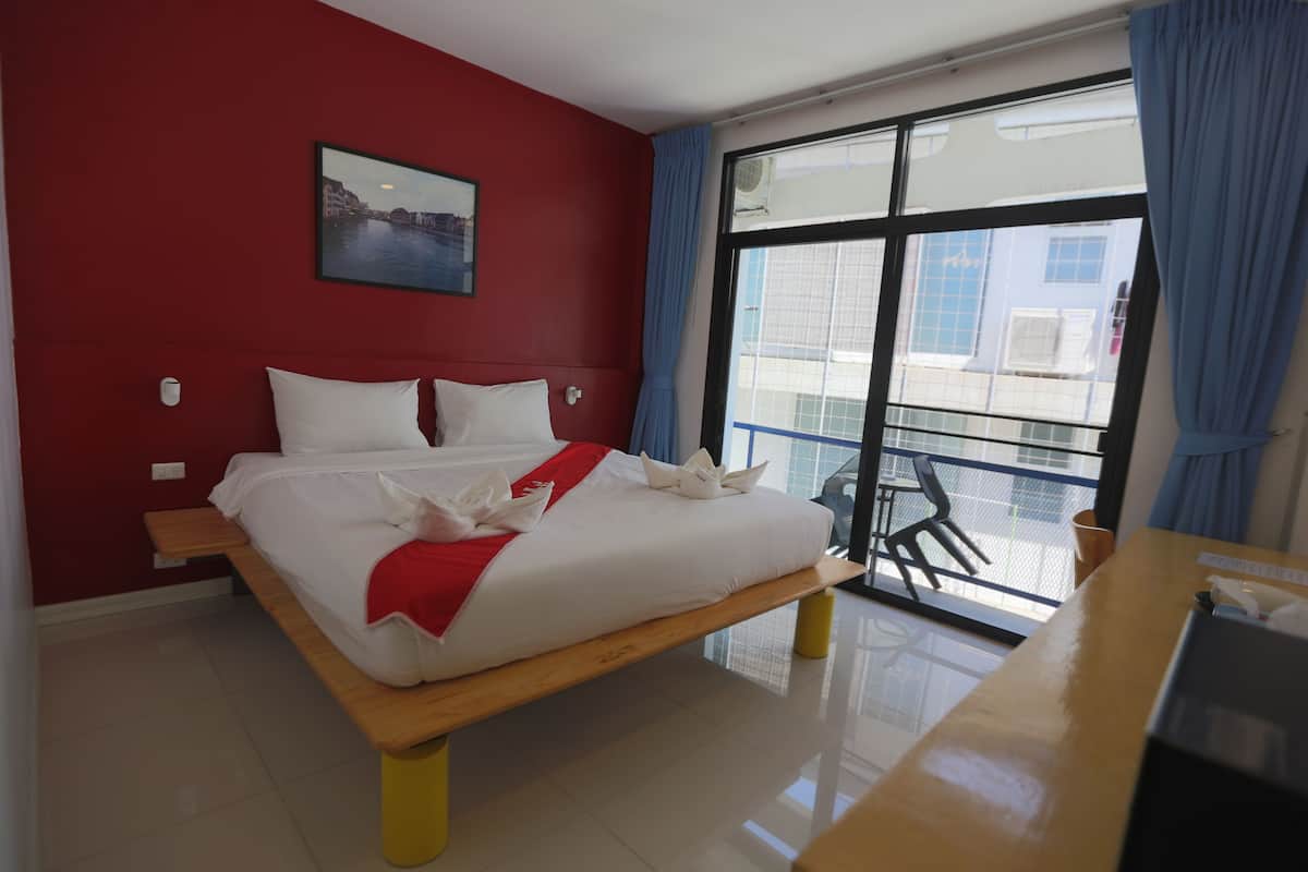 A hotel room with a large bed, a wooden nightstand, a desk, a window with a view of buildings, and a red wall with a picture.