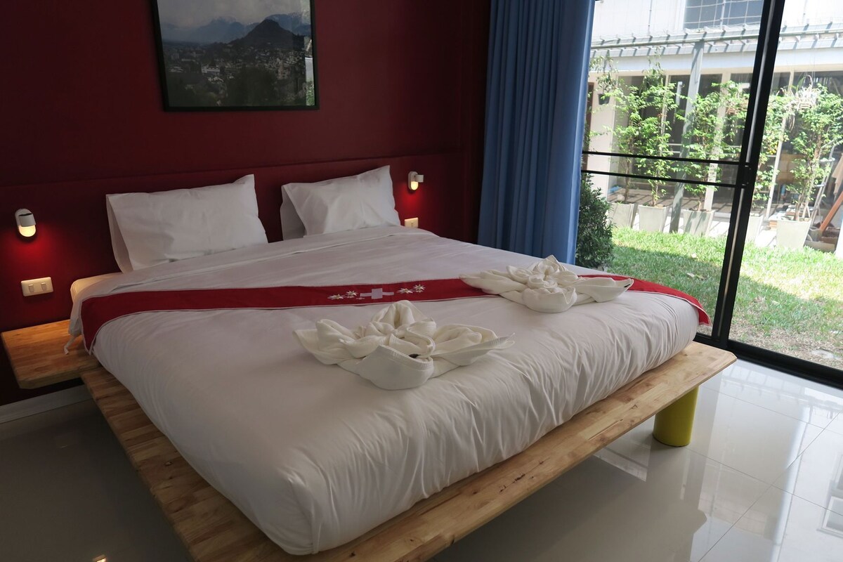 A hotel room with a large bed, a red accent wall, a cityscape view, and a window with a view of greenery.