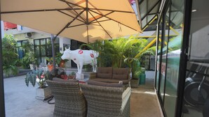 An outdoor seating area with wicker furniture and a large umbrella, surrounded by potted plants and a white cow statue. - Montha Hotel (Chiang Mai)