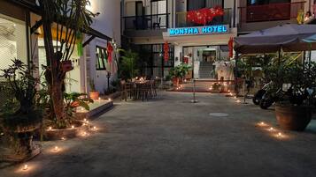 Een hotelentree met een bord waarop 'MONTHA HOTEL' staat, een terras met tafels en stoelen, plantenbakken en een motorfiets voor de deur.
