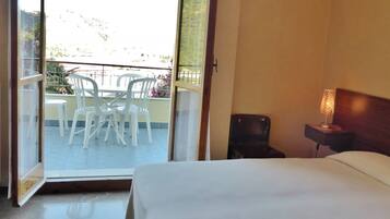 A hotel room with a bed, a nightstand with a lamp, a chair, and a view of an outdoor balcony with chairs and a table.