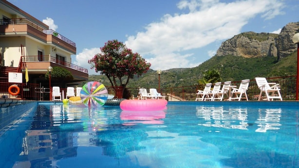 A swimming pool with clear blue water, surrounded by white lounge chairs and a colorful beach ball. A mountain with greenery is visible in the background. - Residence Green Park (Ventimiglia)