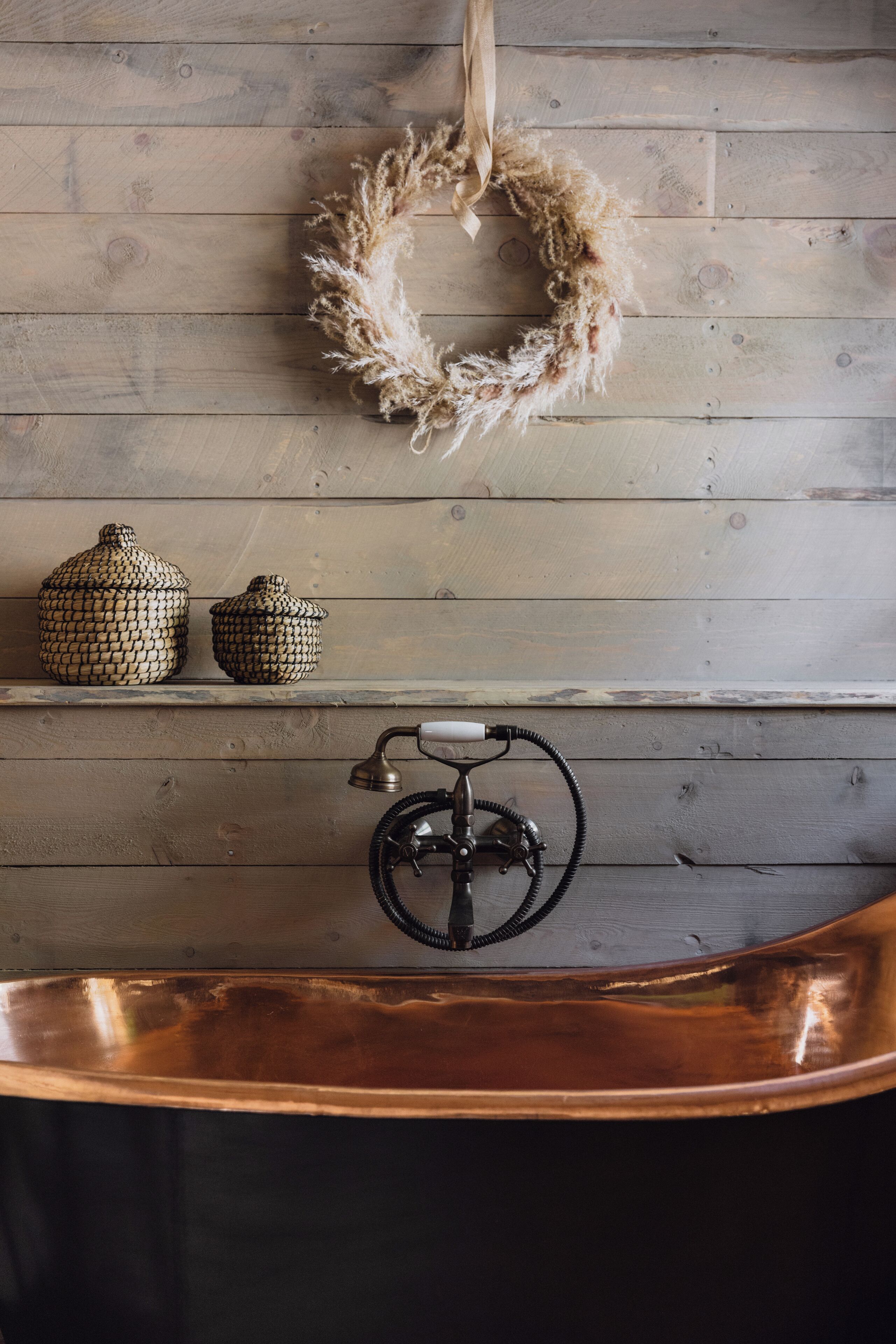 A rustic bathroom with a copper bathtub, a wall-mounted faucet, and two decorative jars on a shelf.