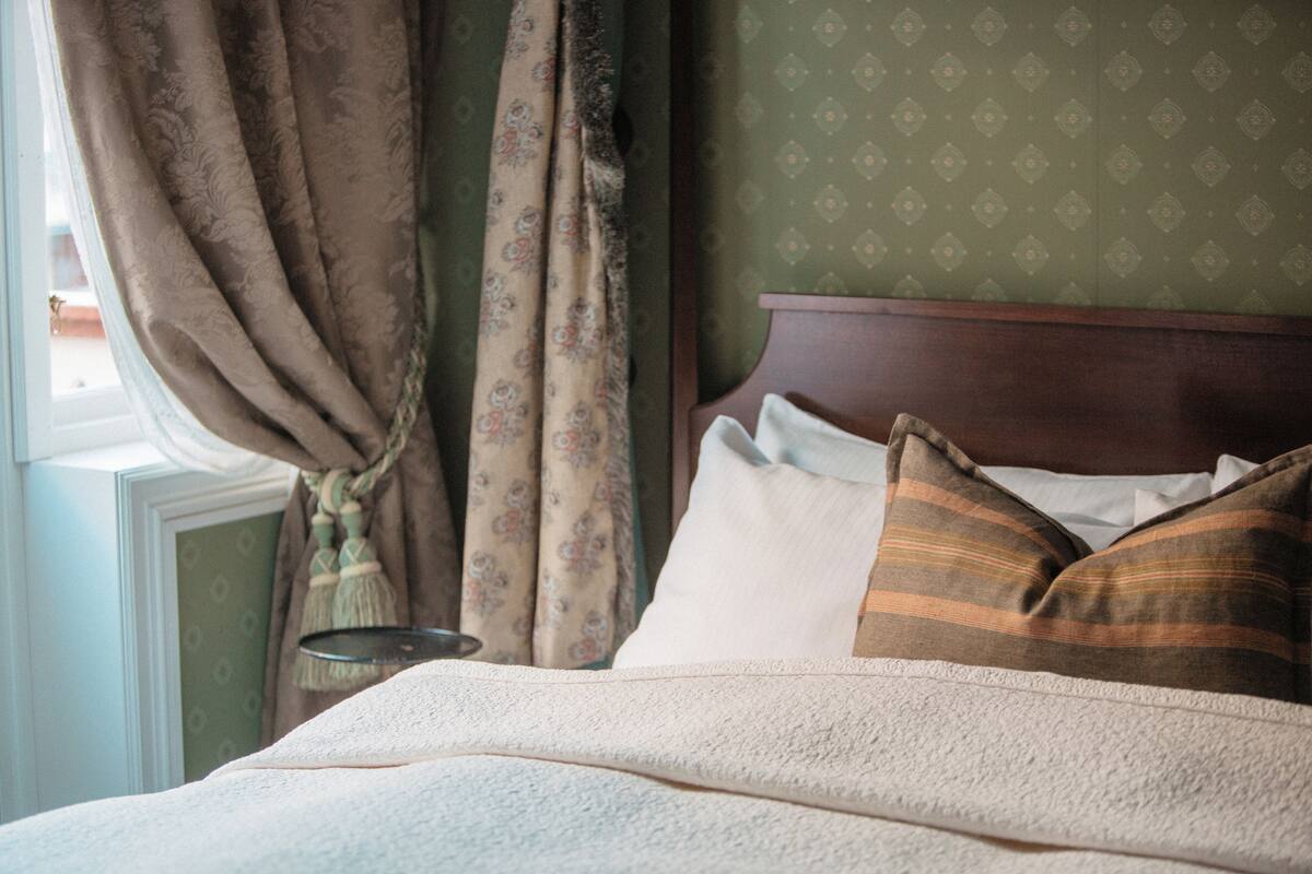 A neatly made bed with a striped pillow and a plain pillow, a wooden headboard, and a window with curtains.