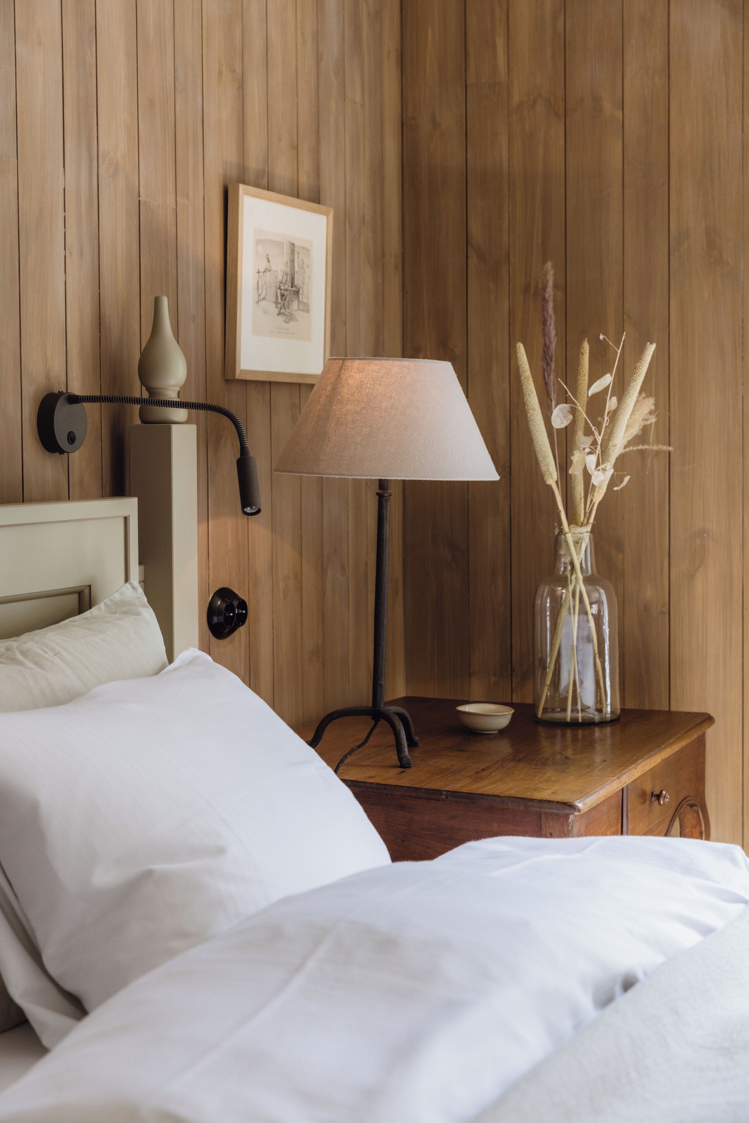 A wooden-paneled room with a bed, a nightstand with a lamp, a framed picture, and a vase with dried plants.