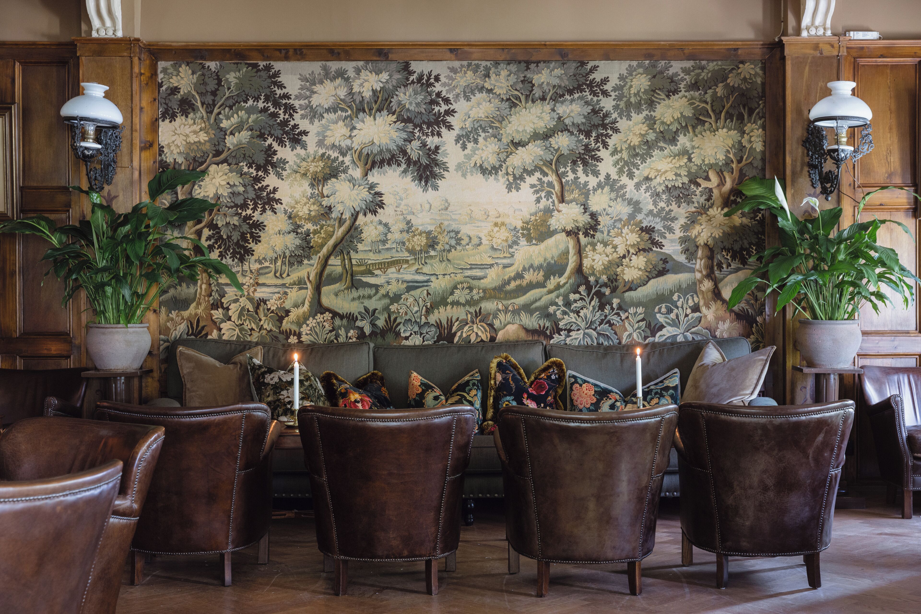 Une pièce comprenant une grande tapisserie représentant un paysage, des fauteuils en cuir, un canapé et deux plantes en pot.