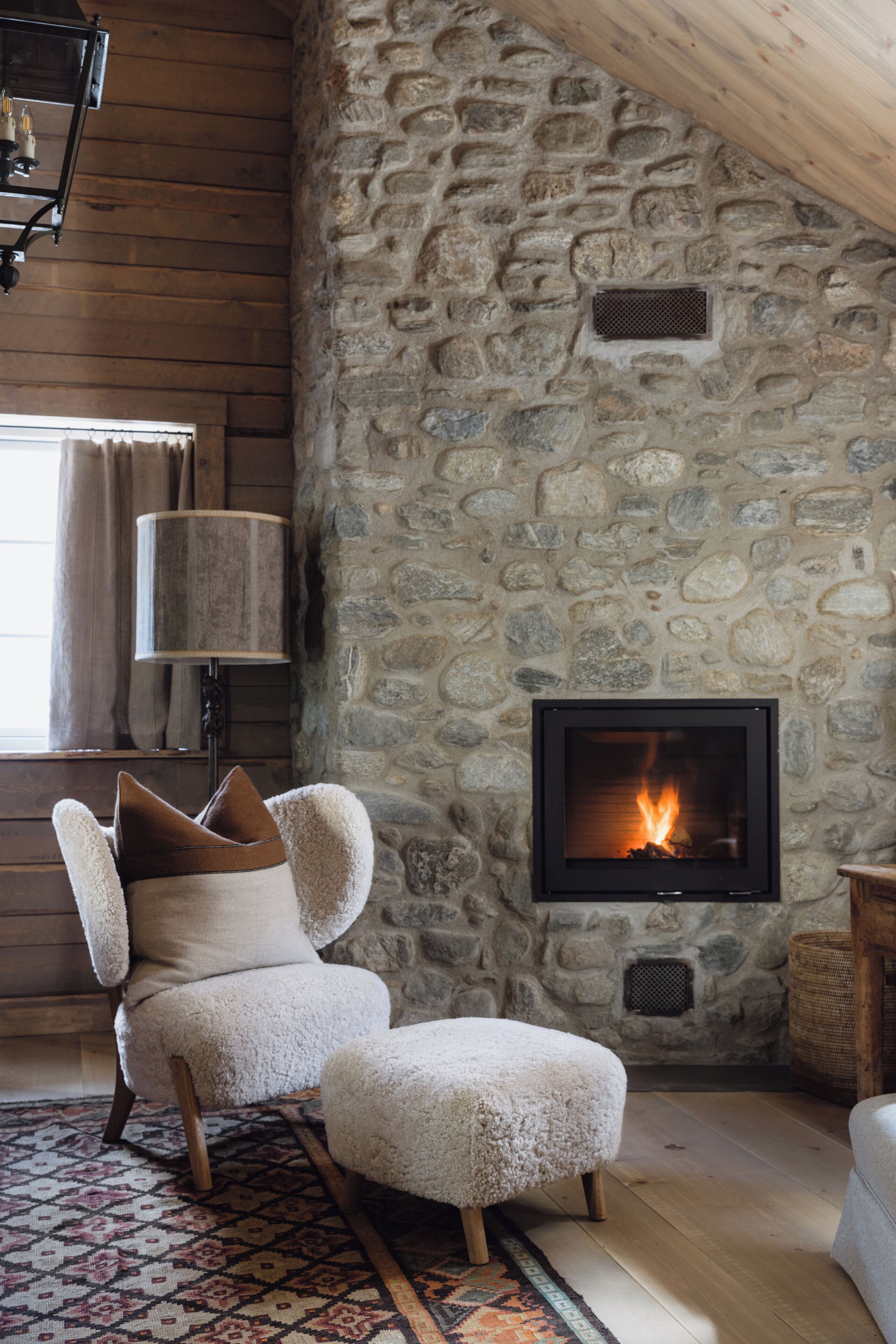 A cozy room with a stone wall, a fireplace, a plush armchair, and a patterned rug.