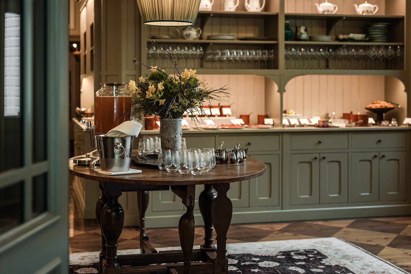A table with a pitcher, glasses, and a vase of flowers in a room with wooden cabinets and a patterned rug.