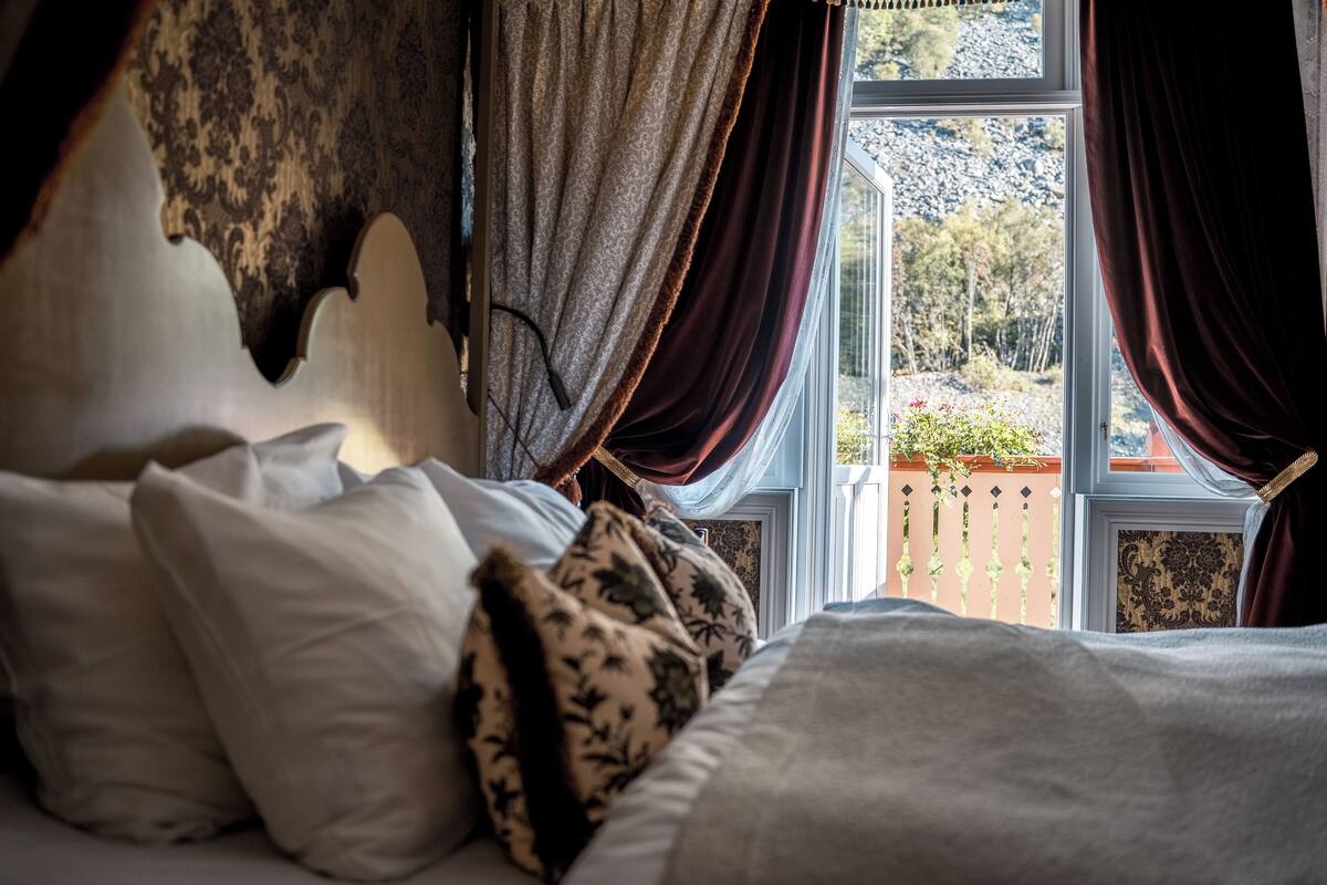 A bed with a headboard, white and patterned pillows, and a view of a garden through a window with curtains.