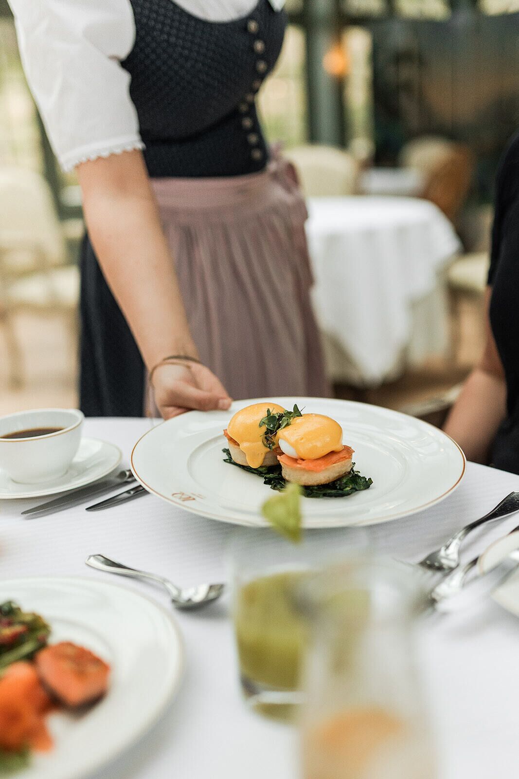 A waiter serving a dish with poached eggs and smoked salmon on a white plate with a gold rim, set on a table with a white tablecloth.