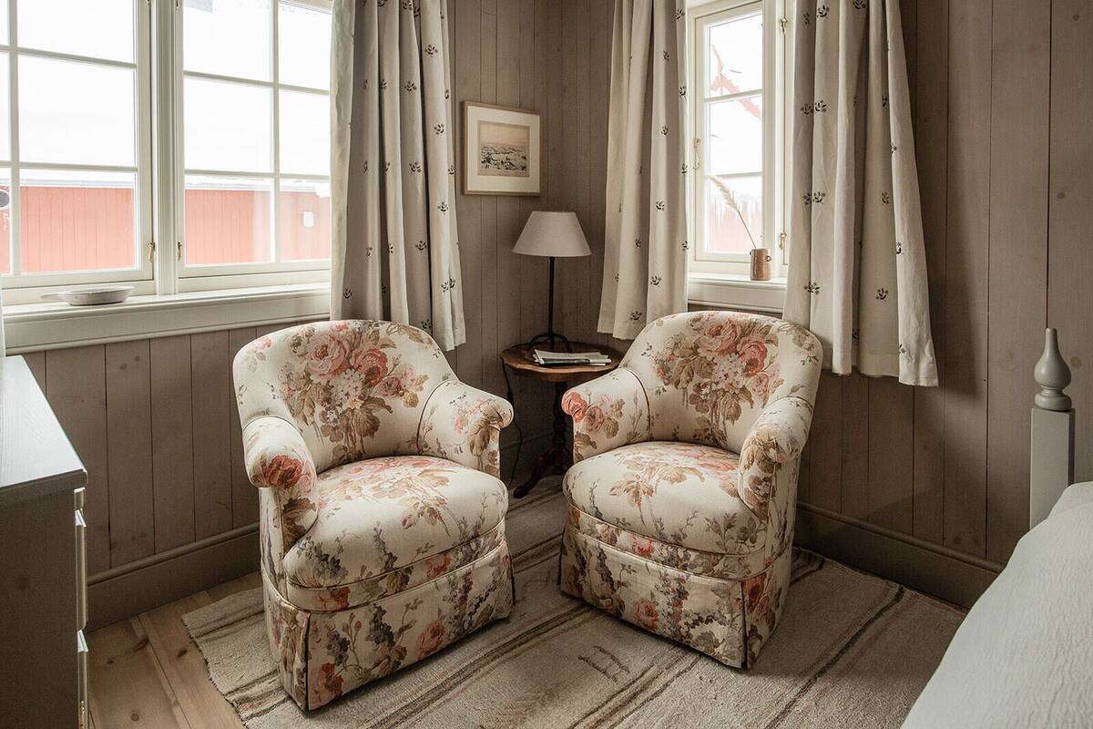 A room with two floral armchairs, a small table, a lamp, and a framed picture on the wall.