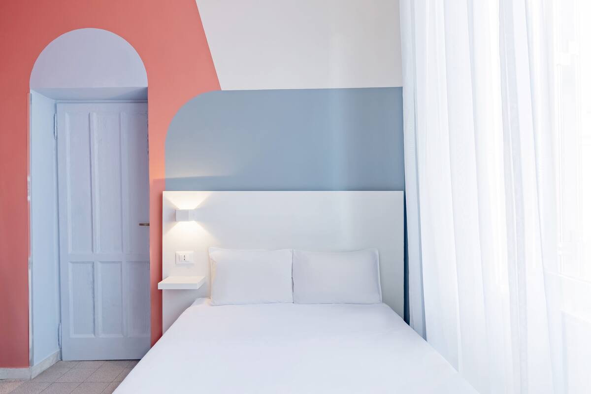 A bedroom with a white bed, a nightstand, a wall-mounted light, and a door.