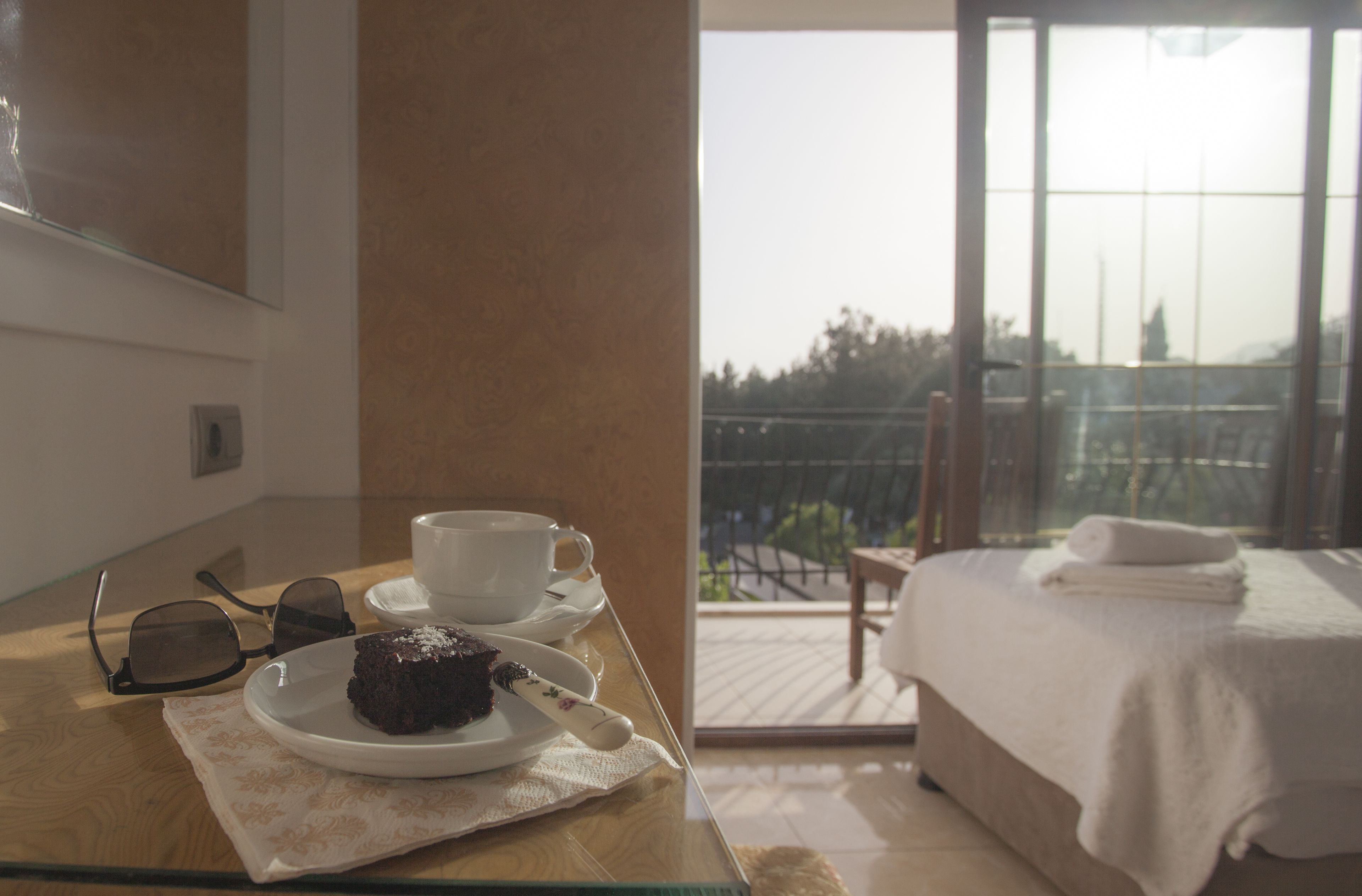 A hotel room with a balcony, a bed with a white cover, a table with a plate of food, a cup, and a pair of glasses.