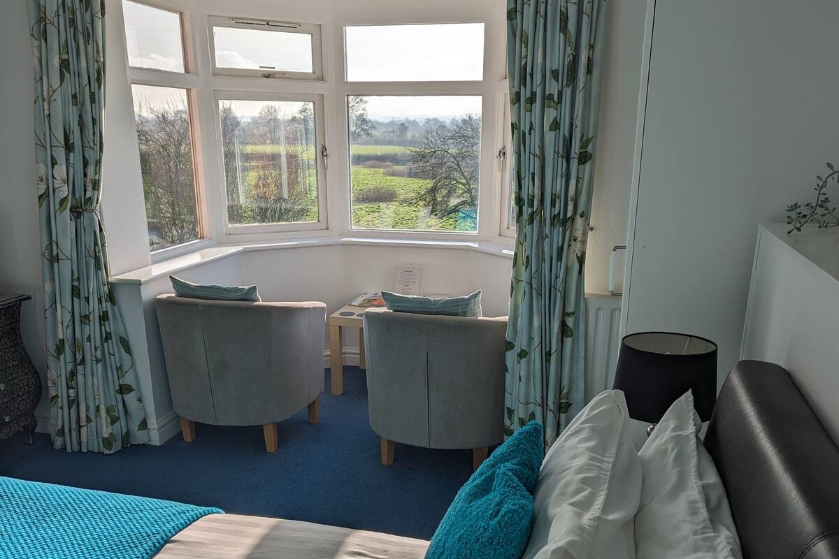 A bedroom with a large bed, a desk, two chairs, and a view of the outdoors through multiple windows.