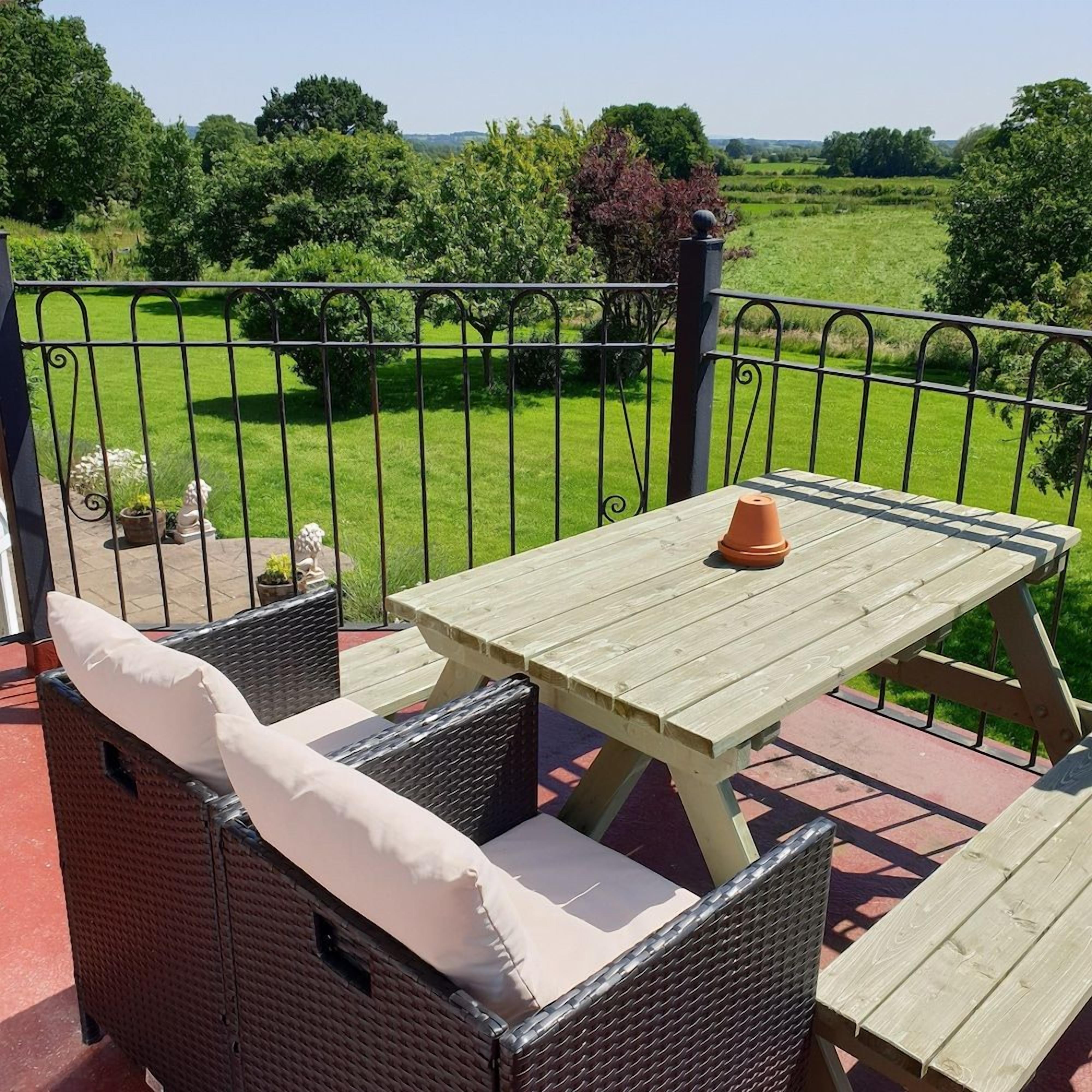 Una mesa de madera y sillas de mimbre en un balcón con barandilla metálica, con vistas a un paisaje verde.