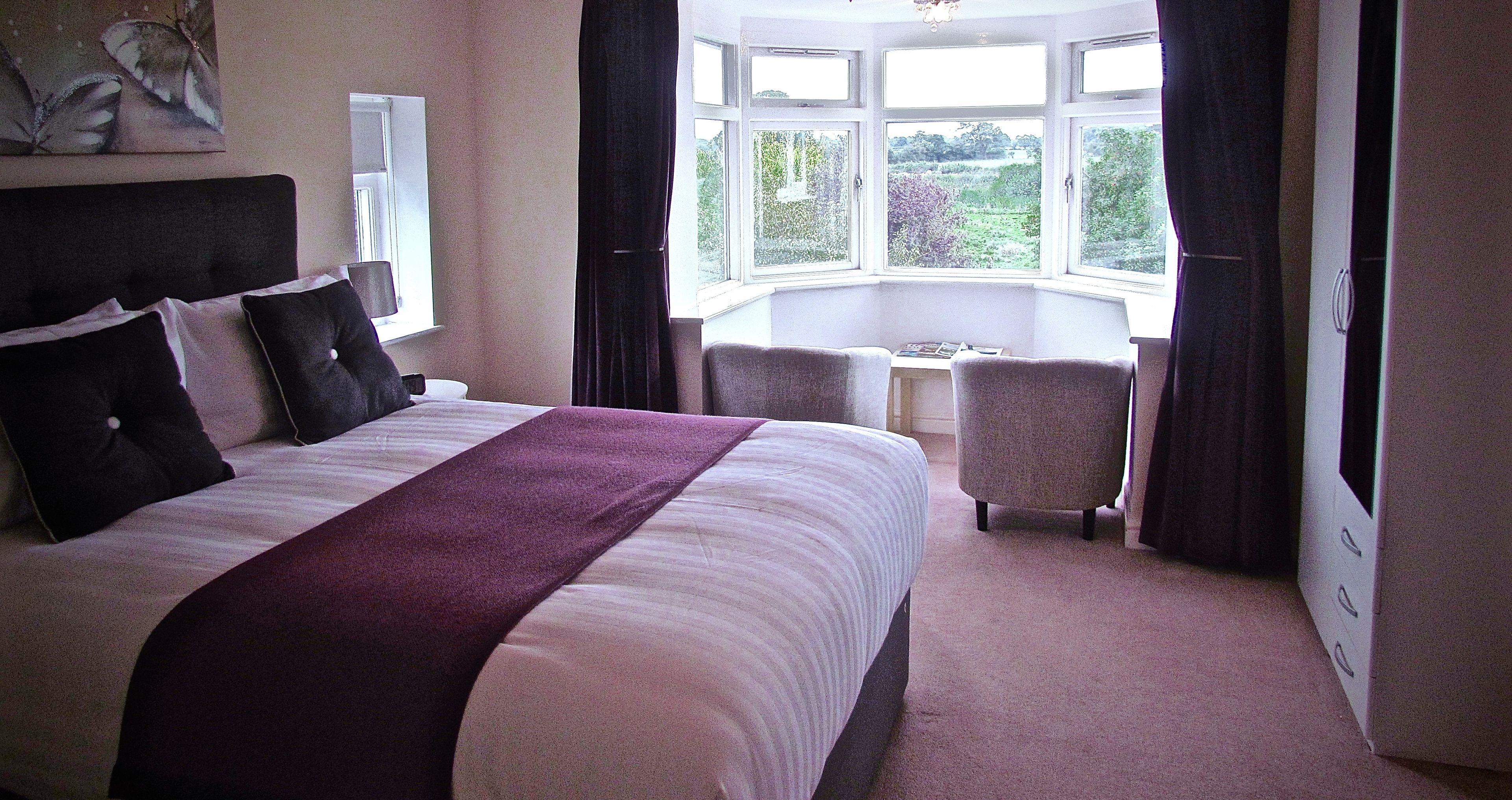 A bedroom with a large bed, a desk, and a view of greenery through the window.