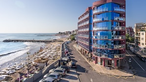 Ett hotell med flera våningar och balkonger, beläget nära en strand med parasoller och bilar.