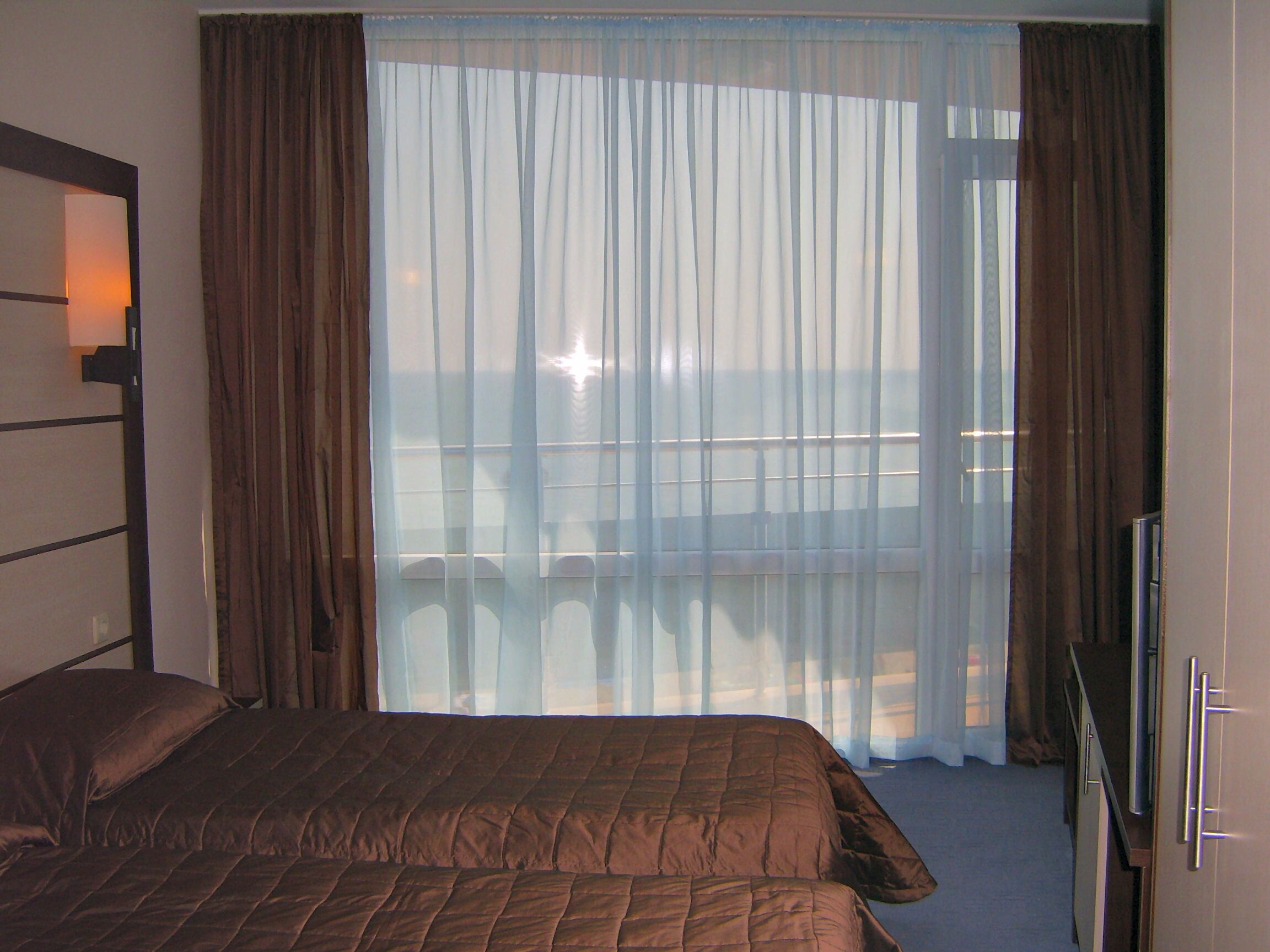 A hotel room with a large bed, a TV, and a view of the sea through sheer curtains.