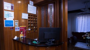 Un mostrador de recepción con una computadora, un teléfono y una lámpara de escritorio. Hay carteles en la pared, incluyendo uno para Wi-Fi y otro referente a un evento de 2013. Se ve una estantería con espacios numerados y una ventana con cortinas al fondo.