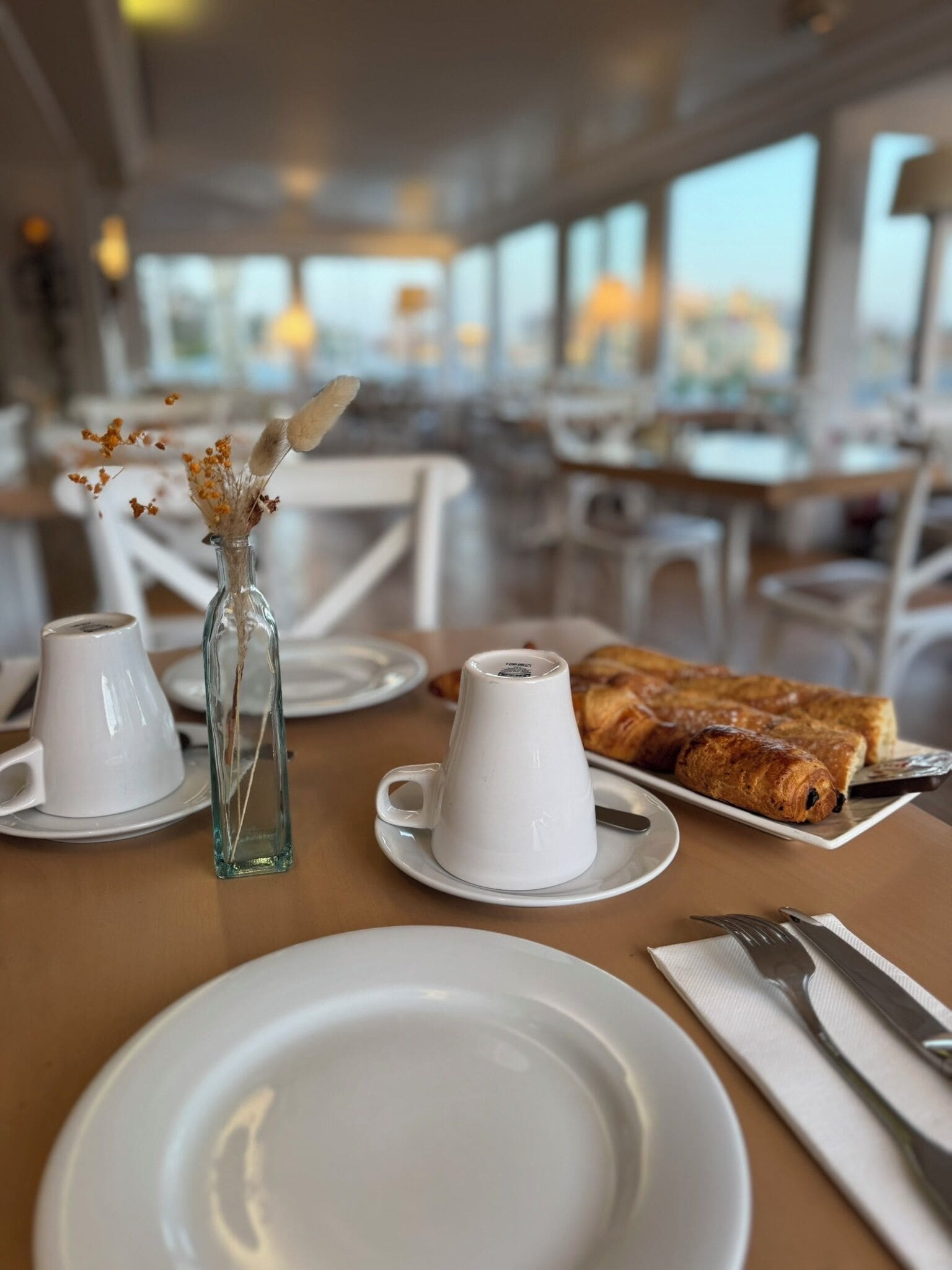A dining table with white plates, a glass vase with dried flowers, and a croissant on a plate.
