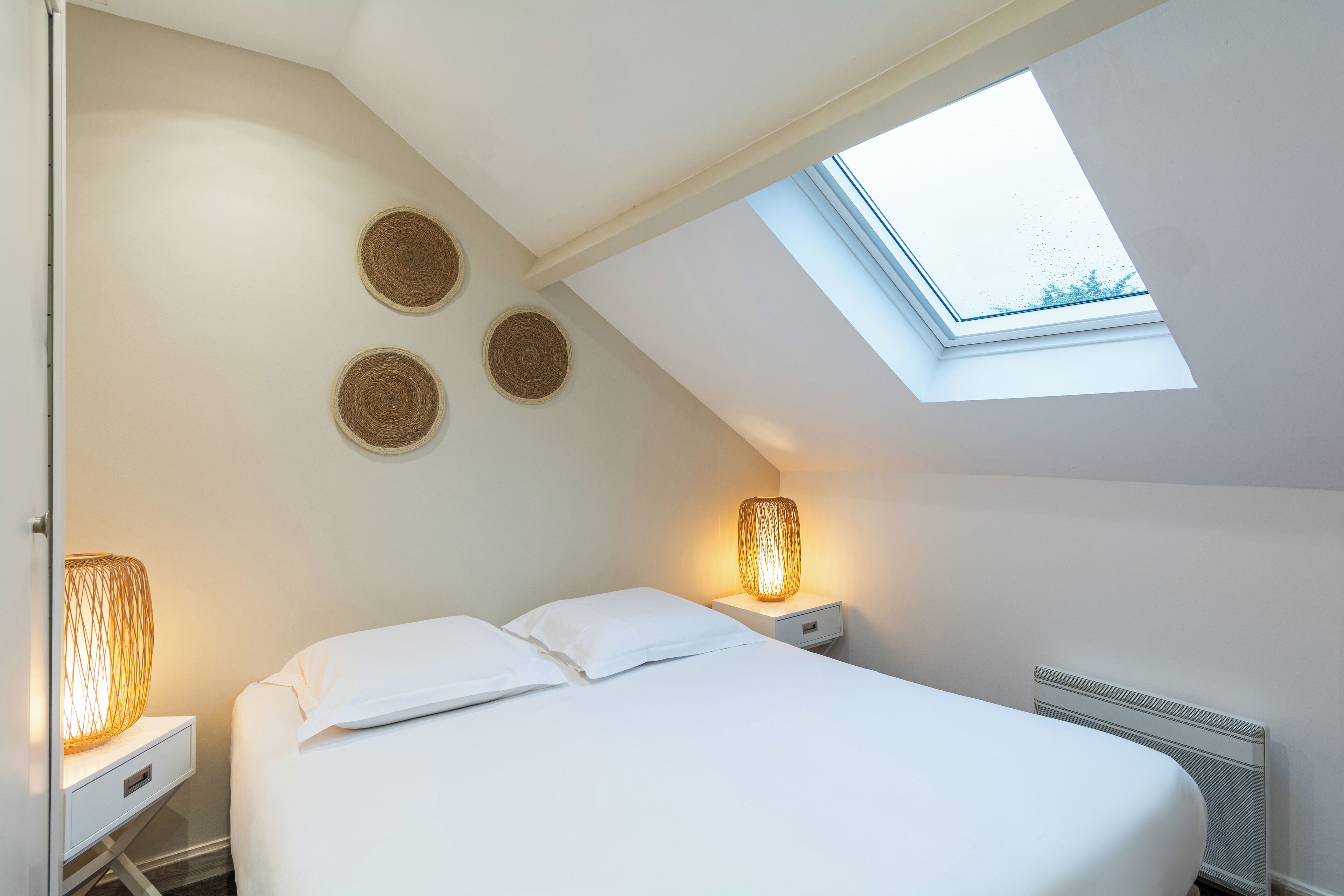 Quarto com claraboia, cama com roupa de cama branca, duas luminárias de apoio e três decorações de parede em tecido.