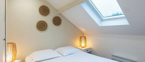 Quarto com claraboia, cama com roupa de cama branca, duas luminárias de apoio e três decorações de parede em tecido.