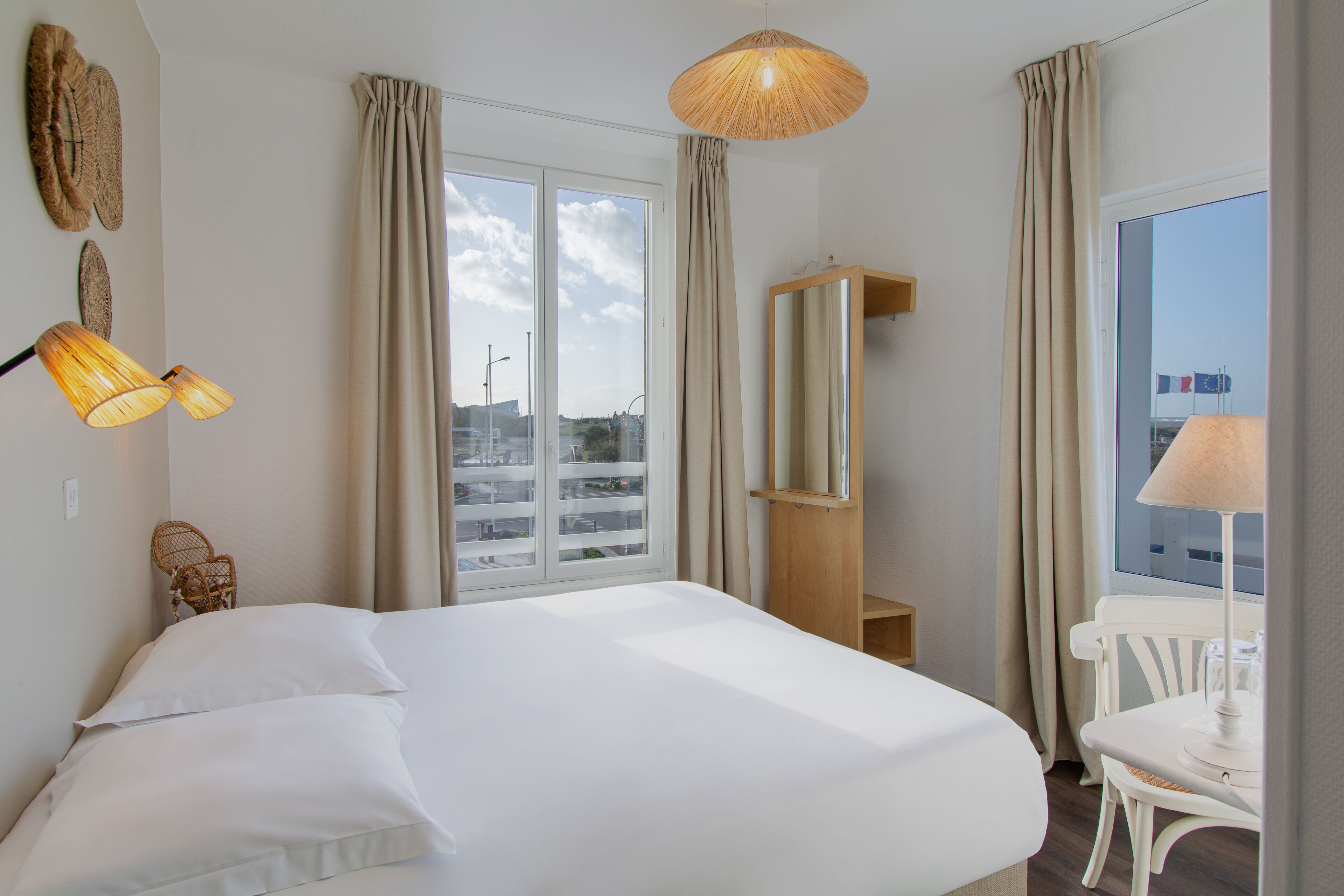A hotel room with a large bed, a chair, a table, and a view of the city through the window.