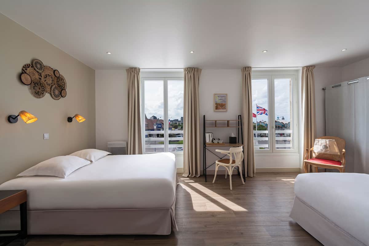 A hotel room with two beds, a desk, a chair, and a view of the city through large windows.