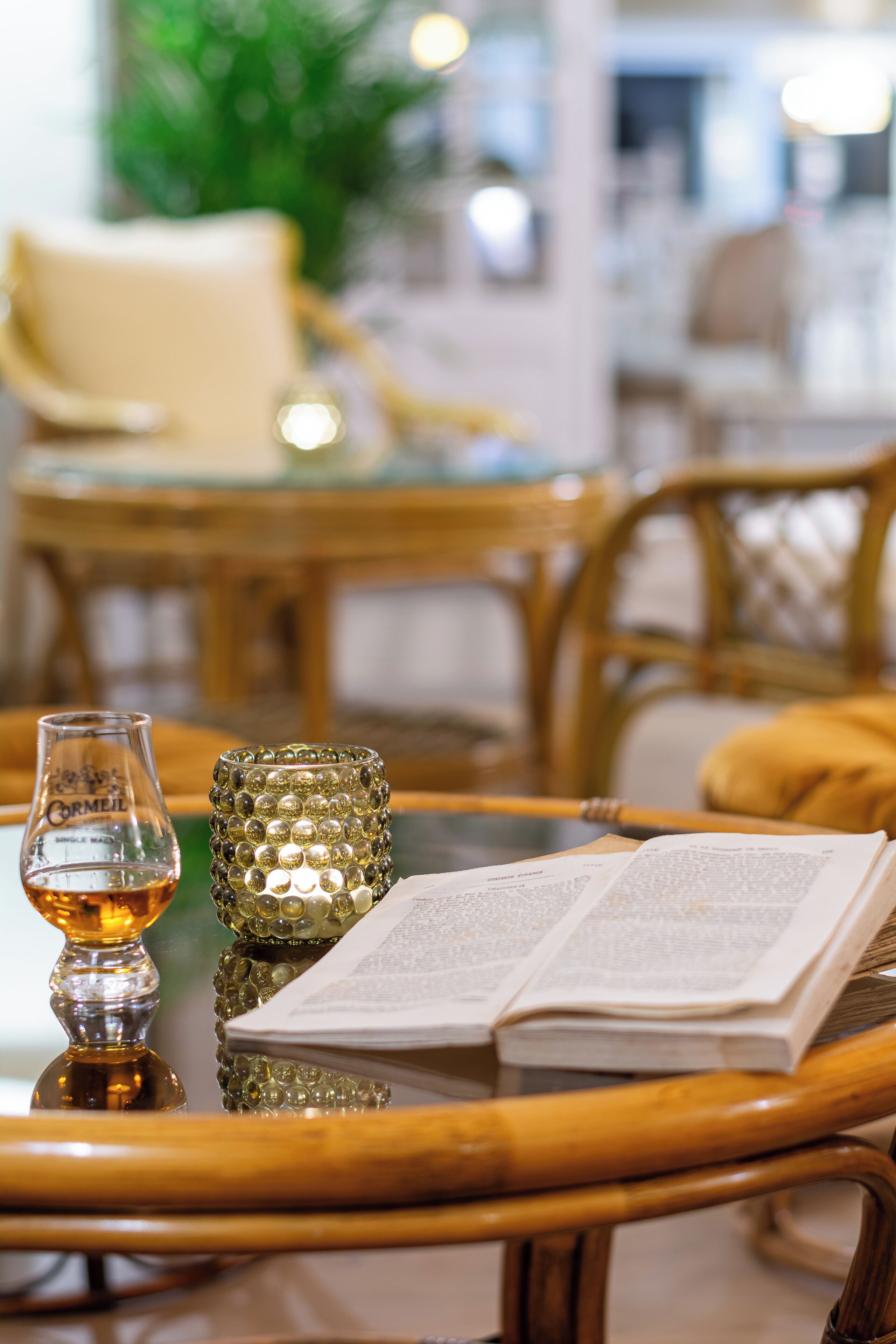 Una silla de mimbre con un cojín, una mesa de vidrio con un libro y una vela, y una botella de vidrio.