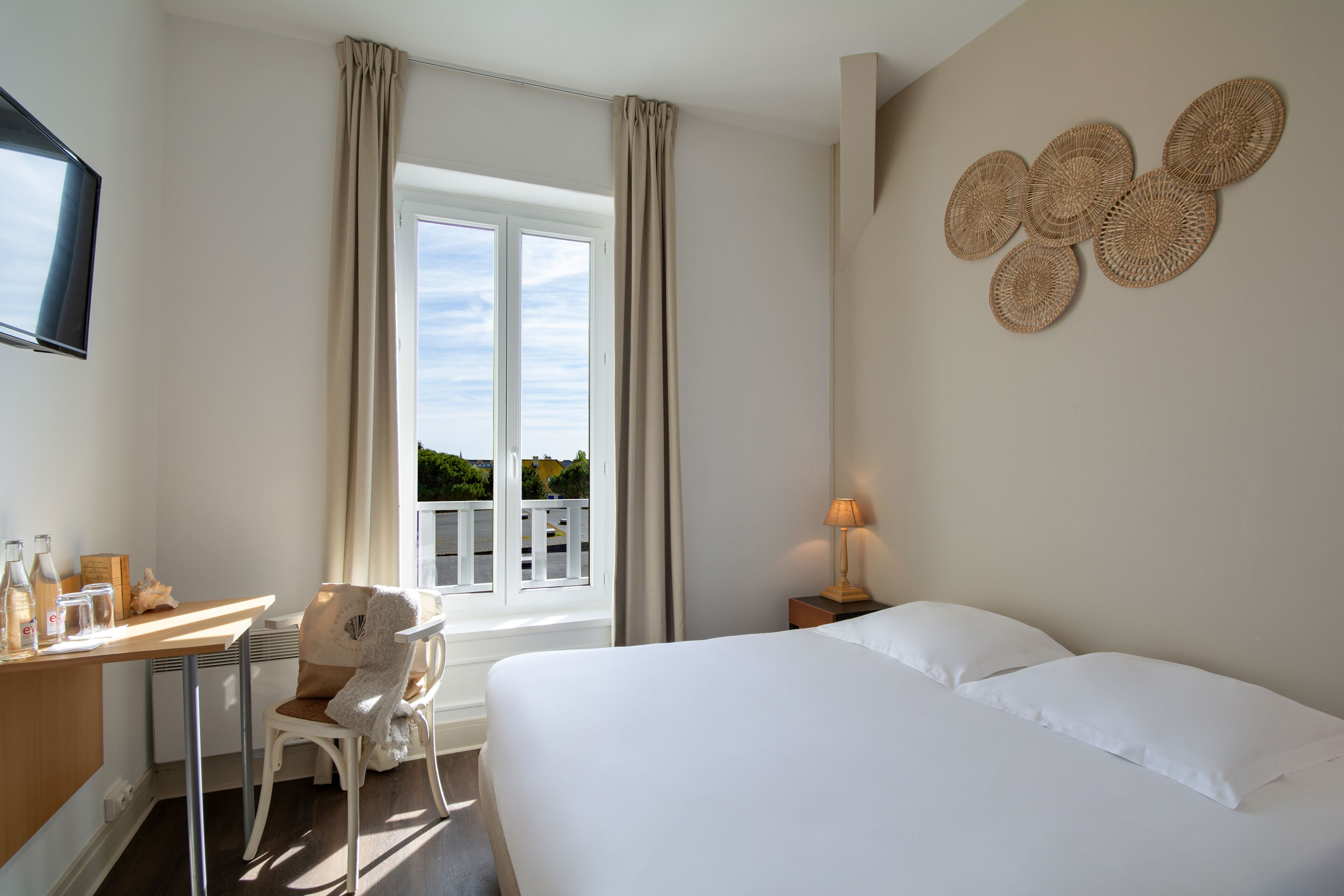 Habitación de hotel con cama, escritorio, silla, televisor, ventana con cortinas y un adorno en la pared.