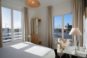 A hotel room with a large bed, a desk with a lamp, a chair, and a view of the city through large windows.