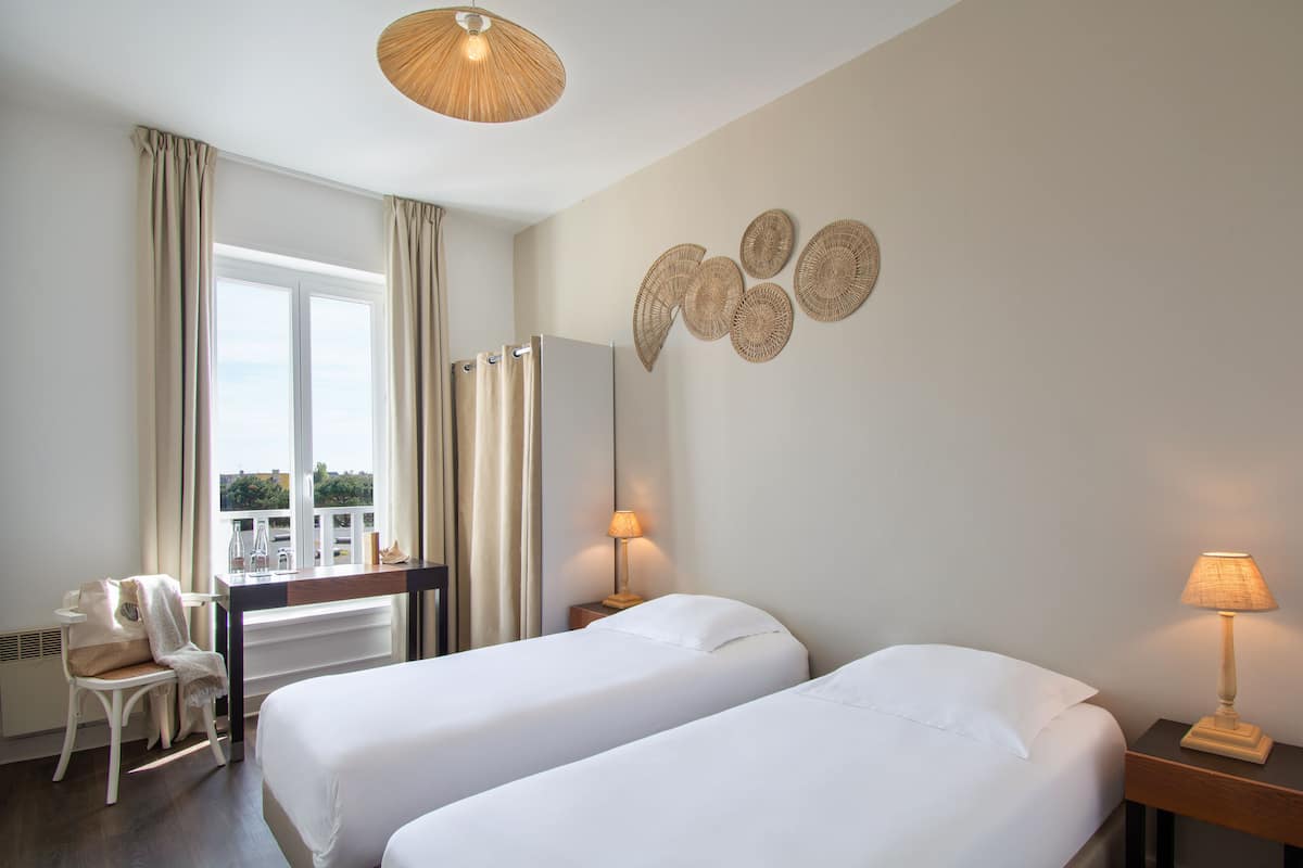 A hotel room with two beds, a desk, a chair, a window with curtains, a wall-mounted lamp, and a ceiling light.