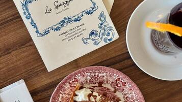 Un menú con diseño floral, un plato con postre, una copa de vino tinto y cubiertos sobre una mesa de madera.