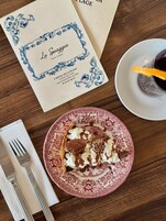 Un menú con diseño floral, un plato con postre, una copa de vino tinto y cubiertos sobre una mesa de madera.
