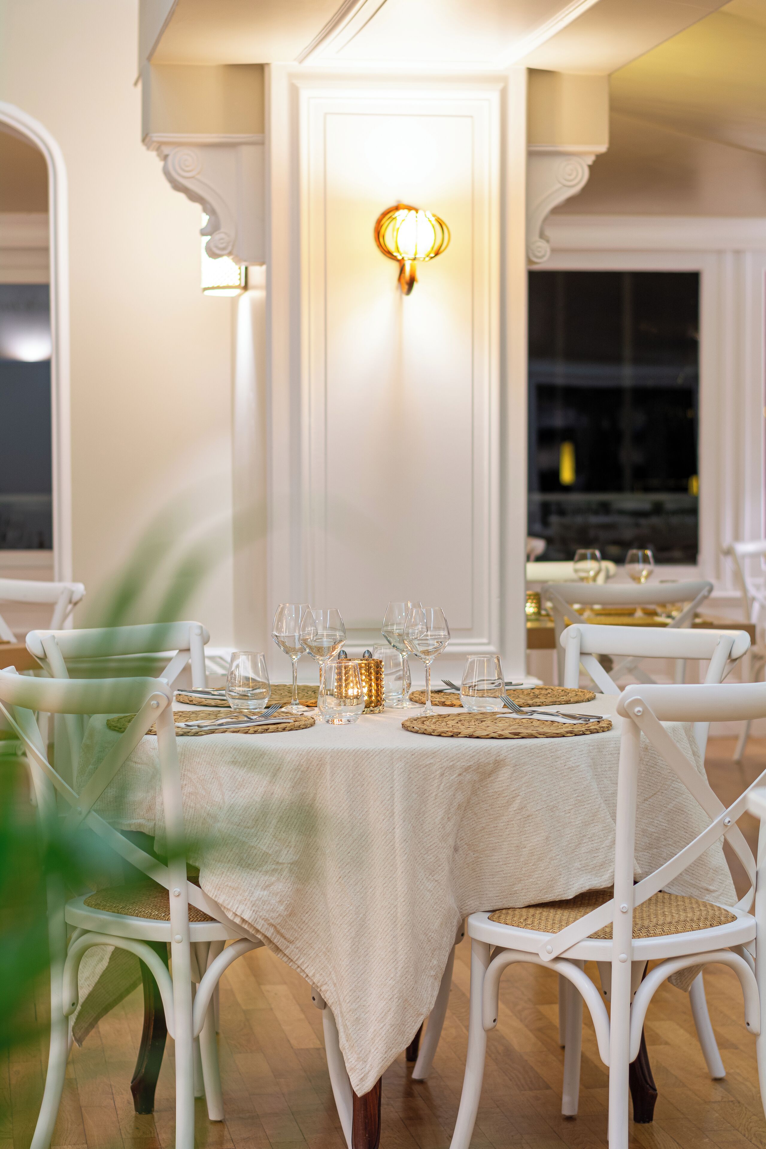 A dining area with white chairs, a table set with wine glasses and plates, and a wall-mounted light fixture.