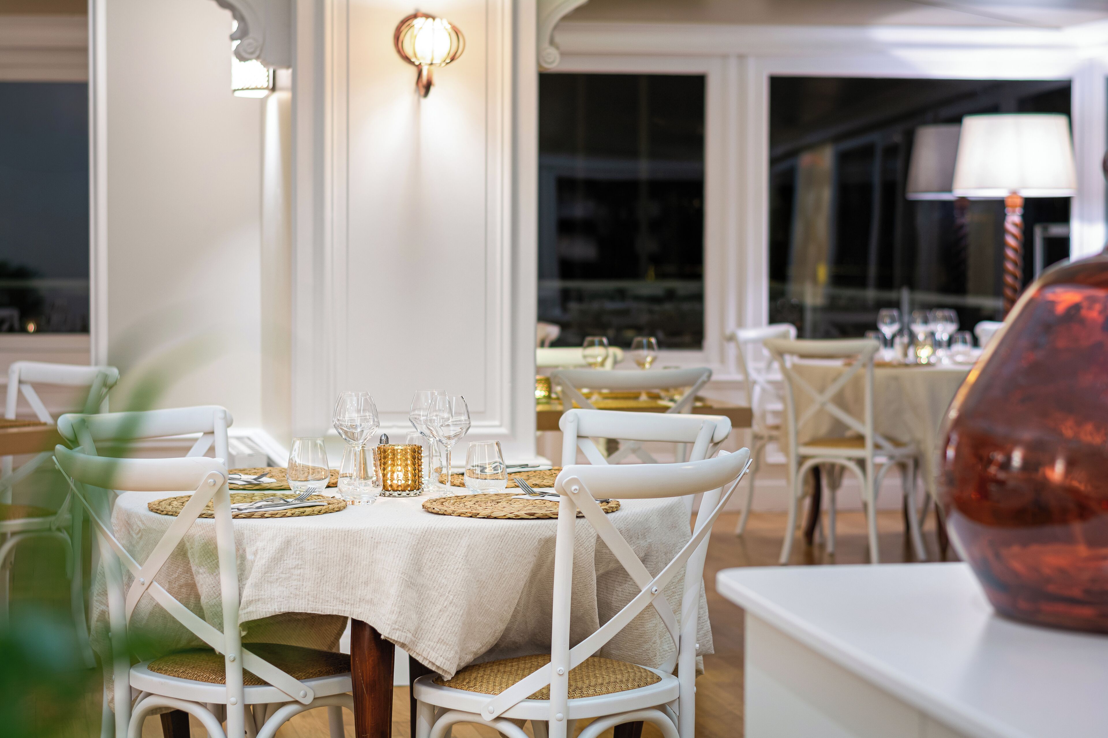 Un espace repas avec des tables rondes disposées pour deux personnes, des chaises blanches et un vase en verre décoratif posé sur une table.