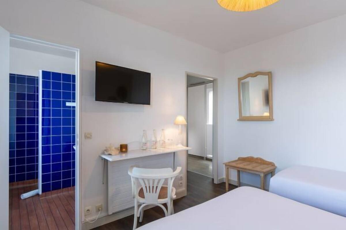 A hotel room with a bed, a desk with a chair, a TV, a mirror, and a bathroom with blue tiles.