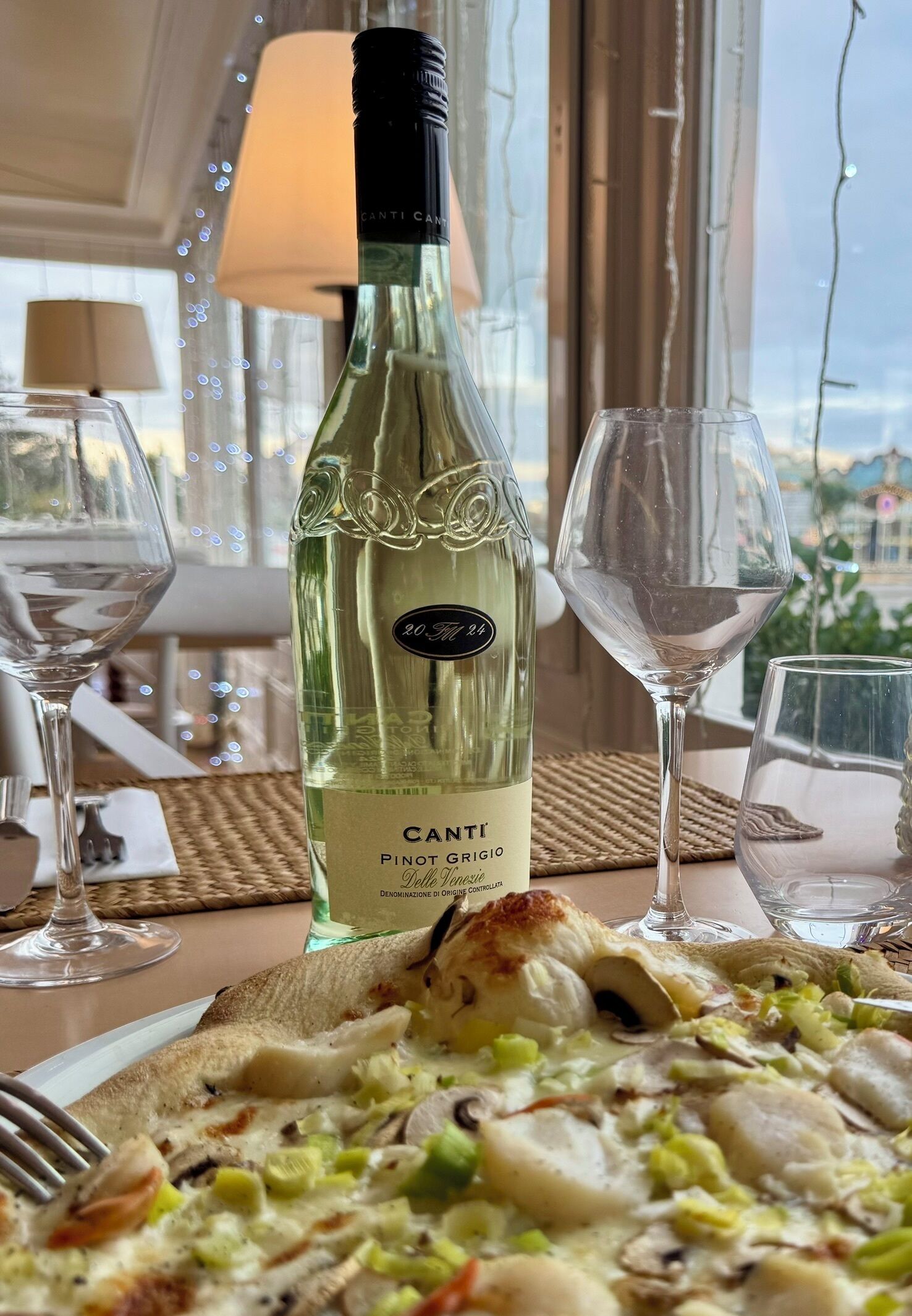 Una botella de vino Canti Pinot Grigio, un plato de comida y dos copas de vino sobre una mesa con vistas.