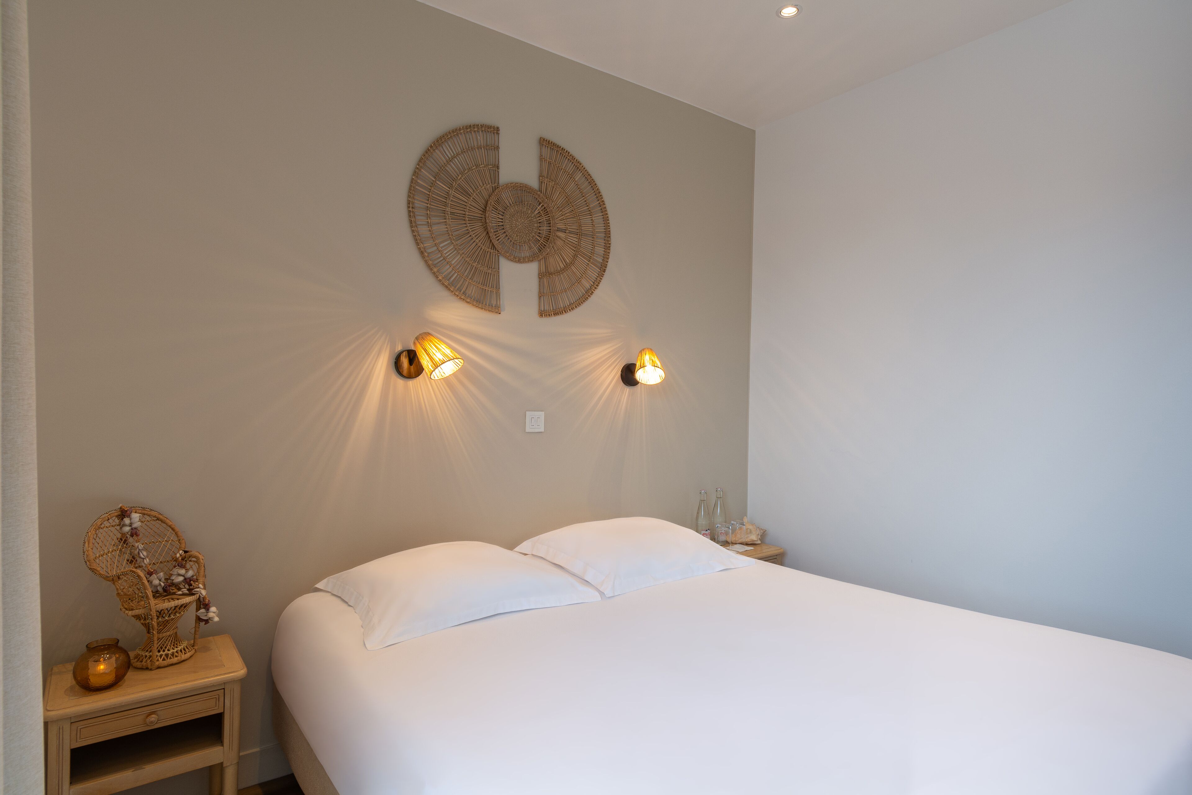Une chambre à coucher avec un lit, deux lampes fixées au mur, un fauteuil décoratif et une petite table sur laquelle sont posées des bouteilles.