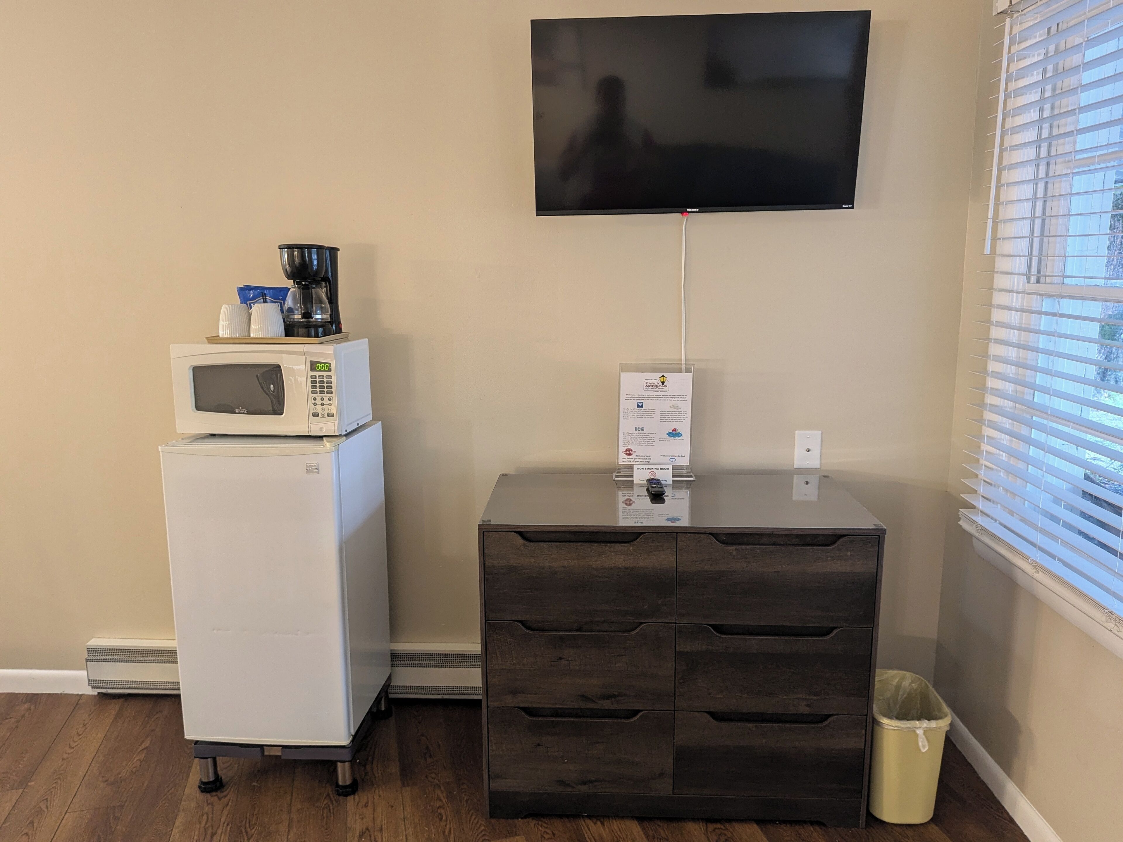A kitchenette with a microwave, coffee maker, and a small refrigerator. There is a TV mounted on the wall and a trash bin next to the counter.