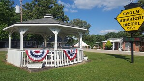 A gazebo with a white fence and American flag decorations, surrounded by a grassy area and a motel sign. - Early American Motel (Hardin)