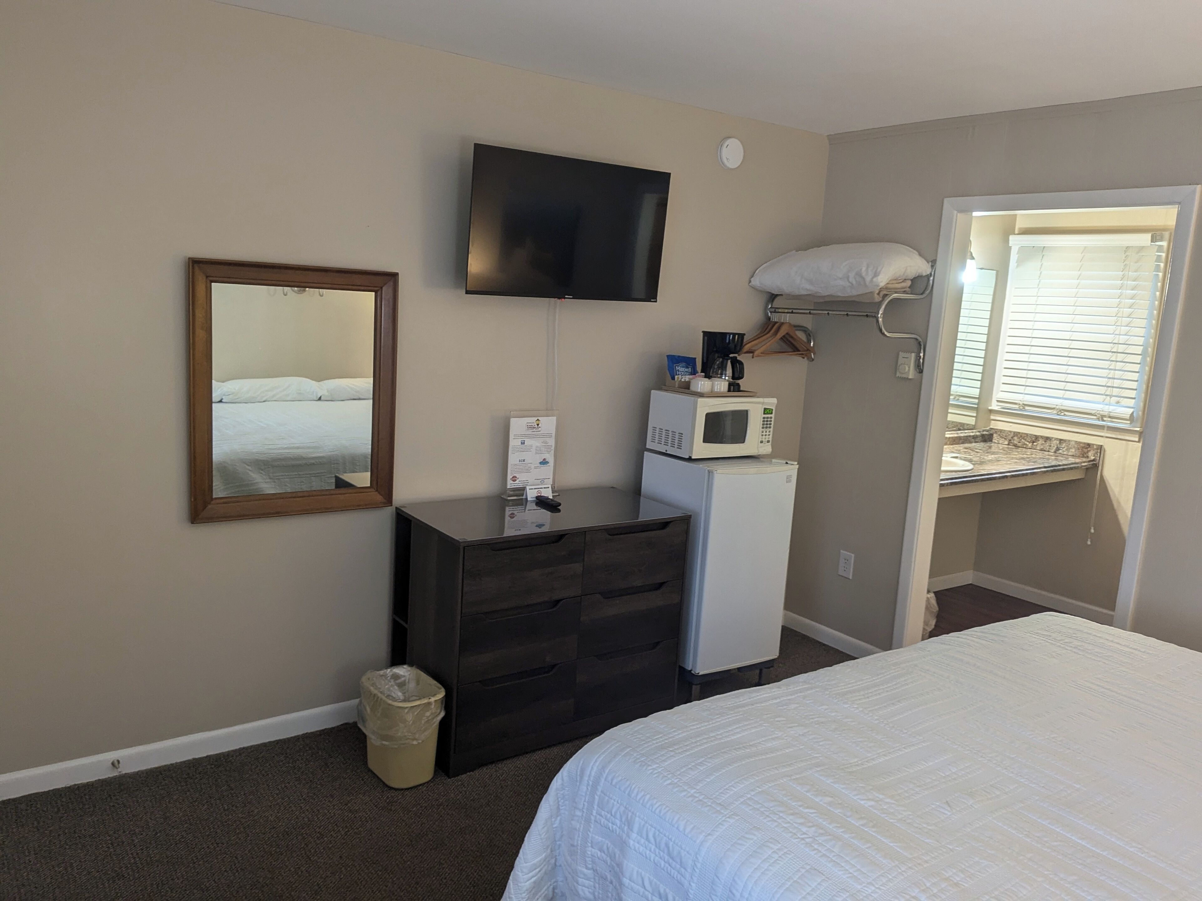A hotel room with a bed, a dresser, a television mounted on the wall, a microwave, and a small kitchenette with a window.