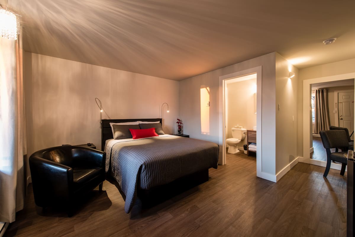 Una habitación de hotel moderna con una cama grande, un sillón de cuero negro, una mesita de noche y un baño con inodoro y lavabo.
